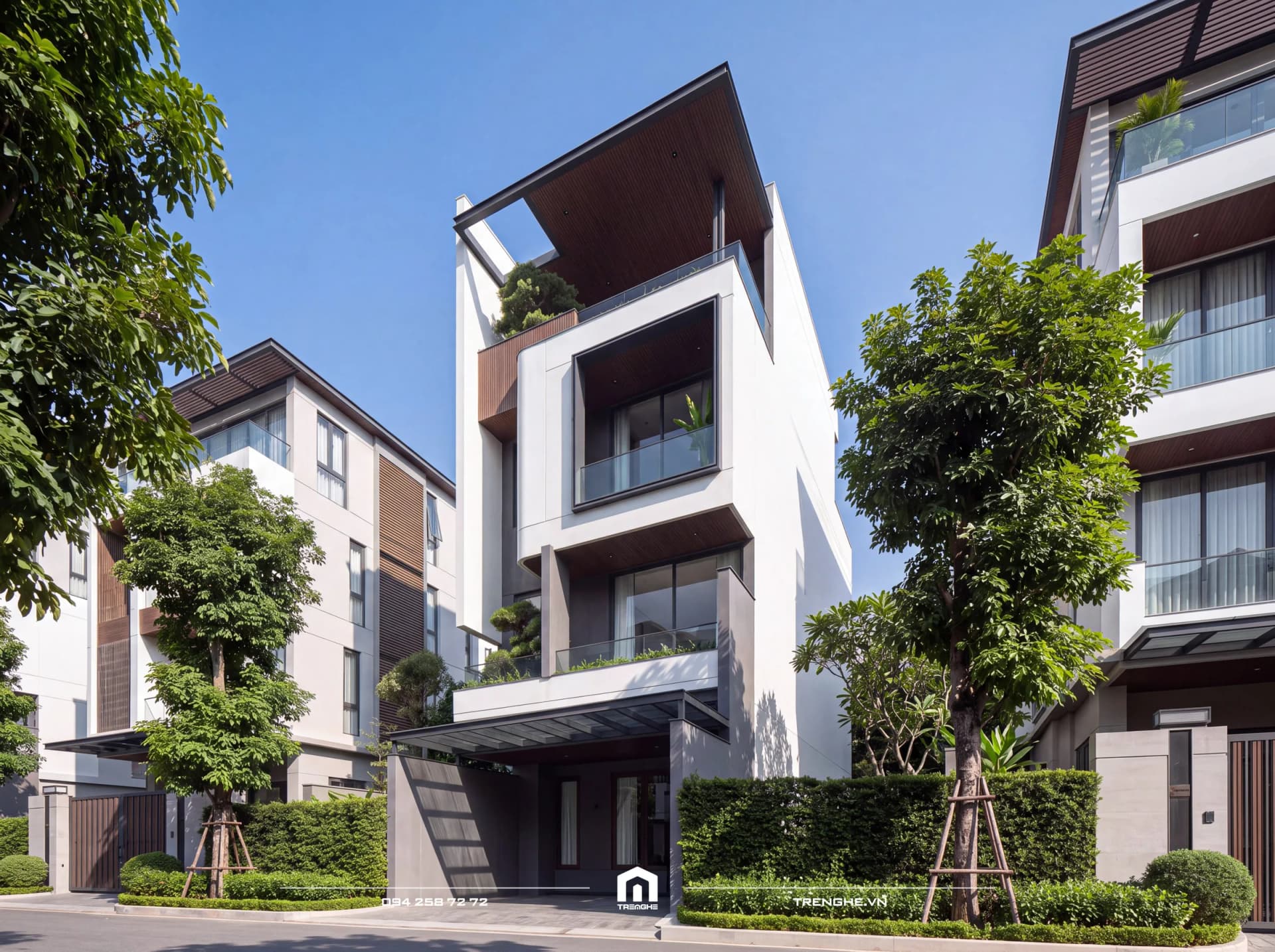 Nhà phố hiện đại T’ House 4 tầng nhìn góc nghiêng: khối tường trắng chữ C ôm ban công kính, mảng ốp gỗ, mái che nghiêng và hàng cây xanh trên phố